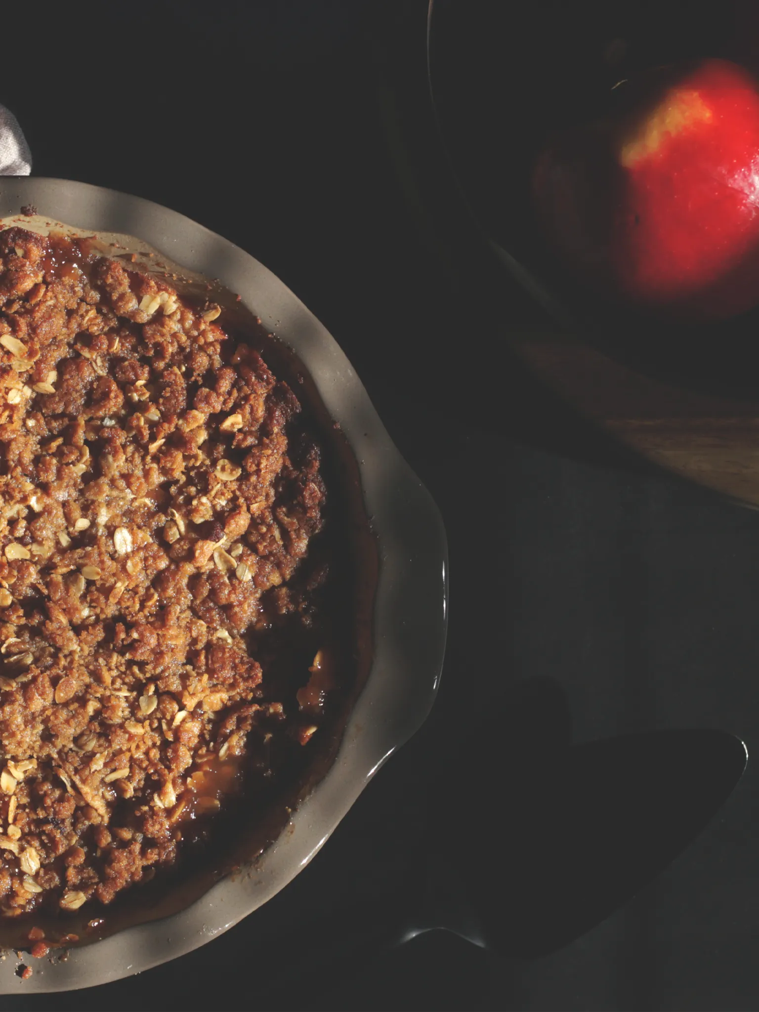 Apple crisp in pie pan next to a red apple on a black table.