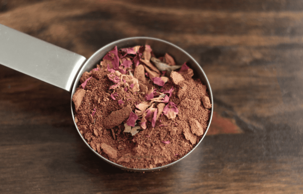 Ground cacao nibs, rose petals, ground cinnamon, and ground cardamom in a measuring cup