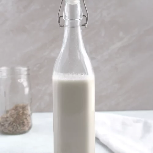 A glass container of homemade oat milk with dried oat seed in the background.