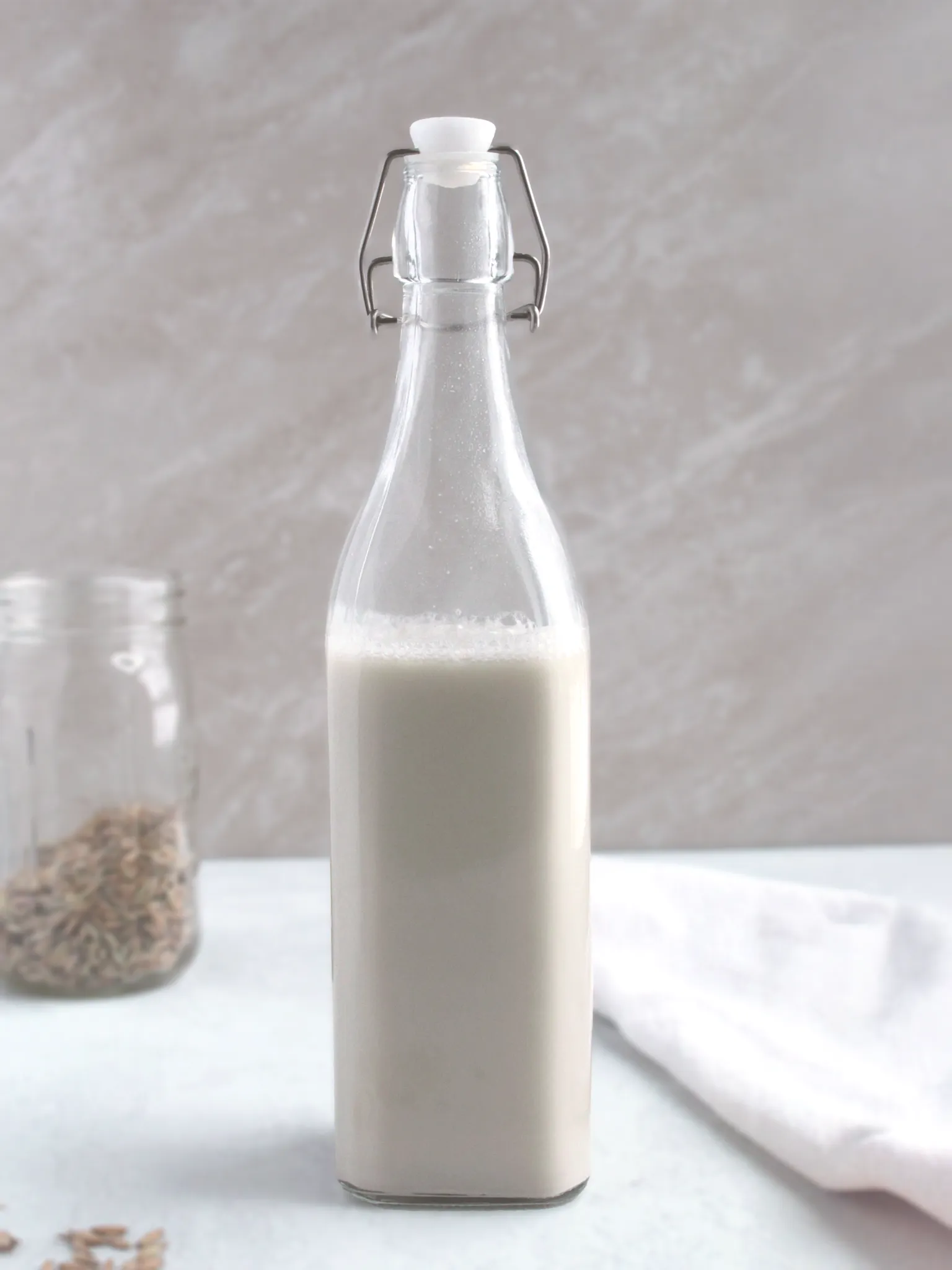 A glass container of homemade oat milk with dried oat seed in the background.