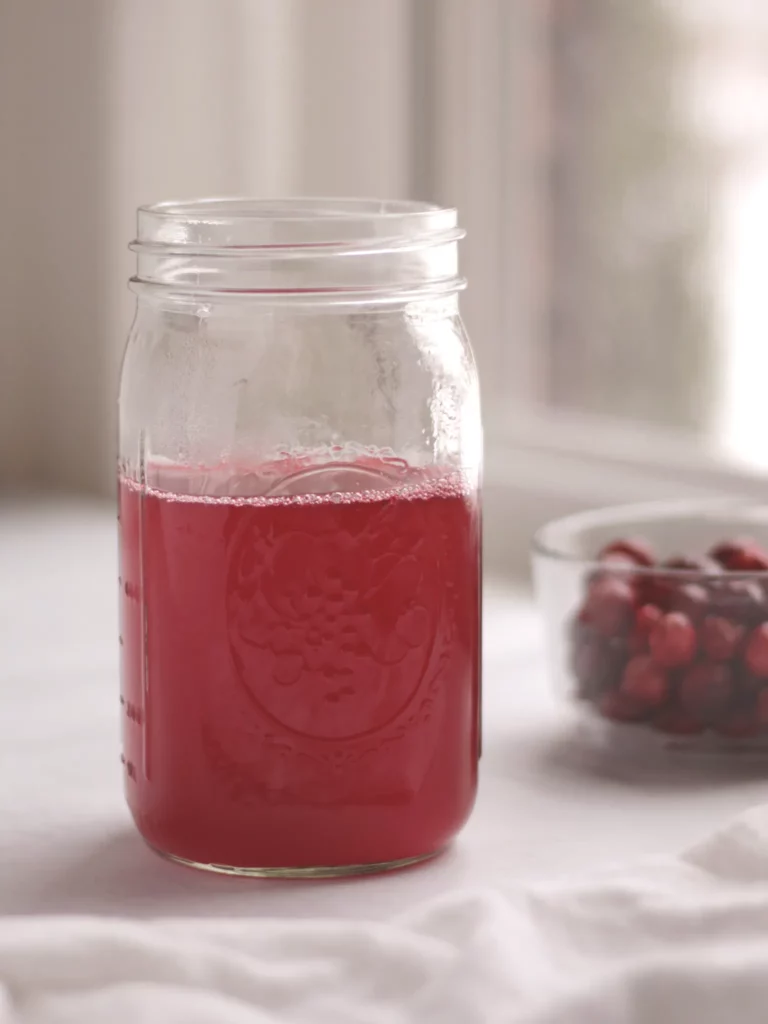 Mason jar filled with homemade cranberry juice on a table next to a bowl of fresh cranberries.