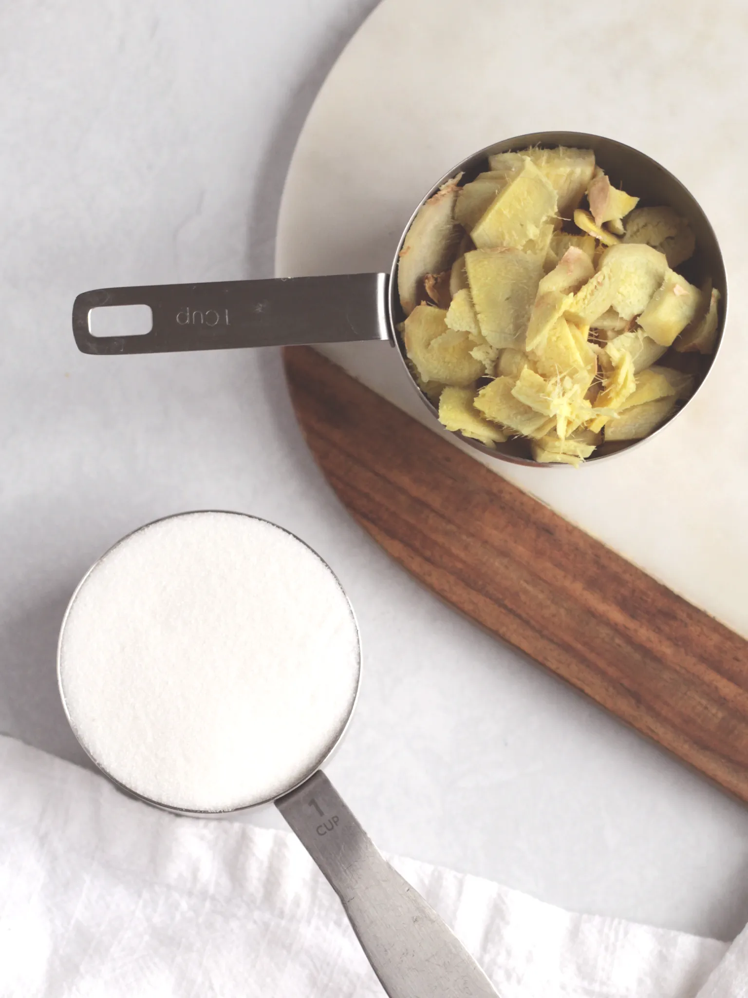 Sliced ginger in a measuring cup next to white sugar in a measuring cup