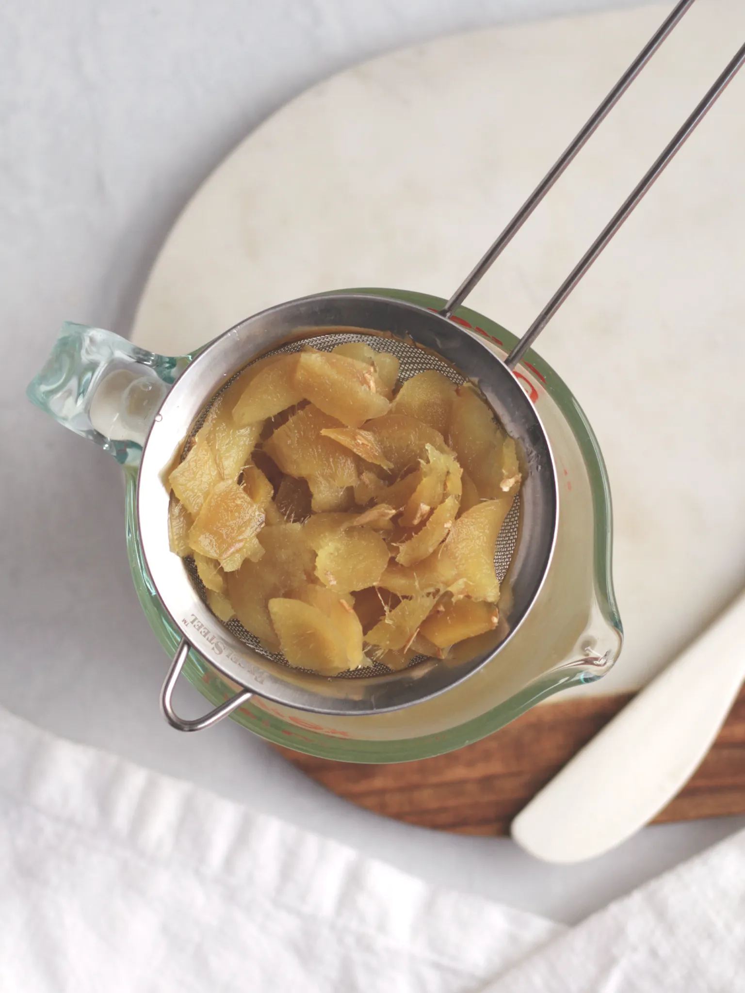 Cooked ginger slices in a fine mesh strainer over a glass measuring cup holding ginger syrup