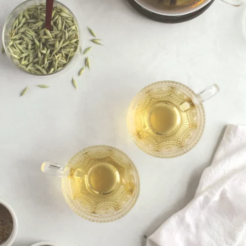 Lemon verbena tea in 2 glass cups with dried oat tops, dried chamomile, and dried lemon verbena on a table.