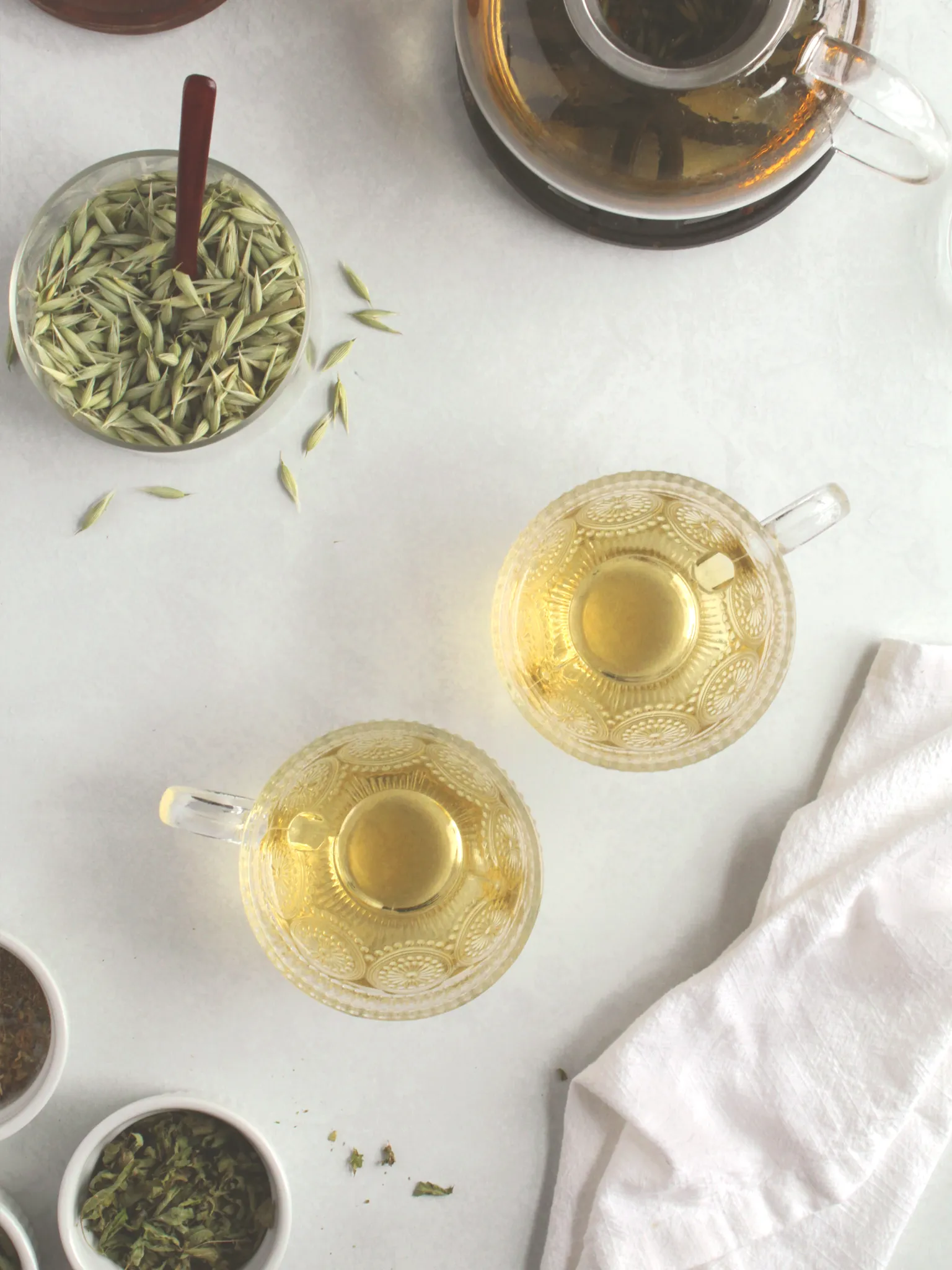 Lemon verbena tea in 2 glass cups with dried oat tops, dried chamomile, and dried lemon verbena on a table.