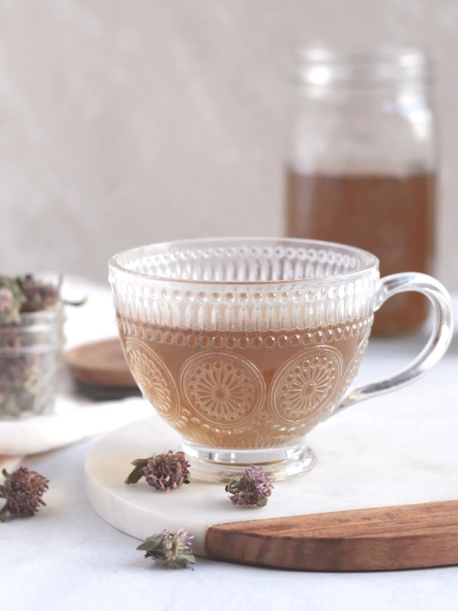 Red clover tea in a glass cup on a table with dried red clover flowers and more tea in a Mason jar.