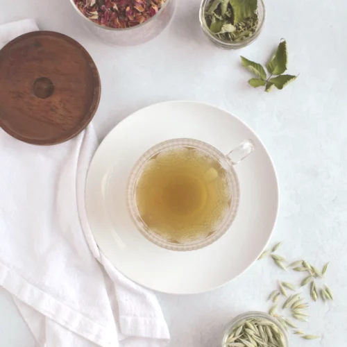 A glass cup of rose blackberry tea on a plate surrounded by containers of dried roses, backberry leaves, and oat tops.