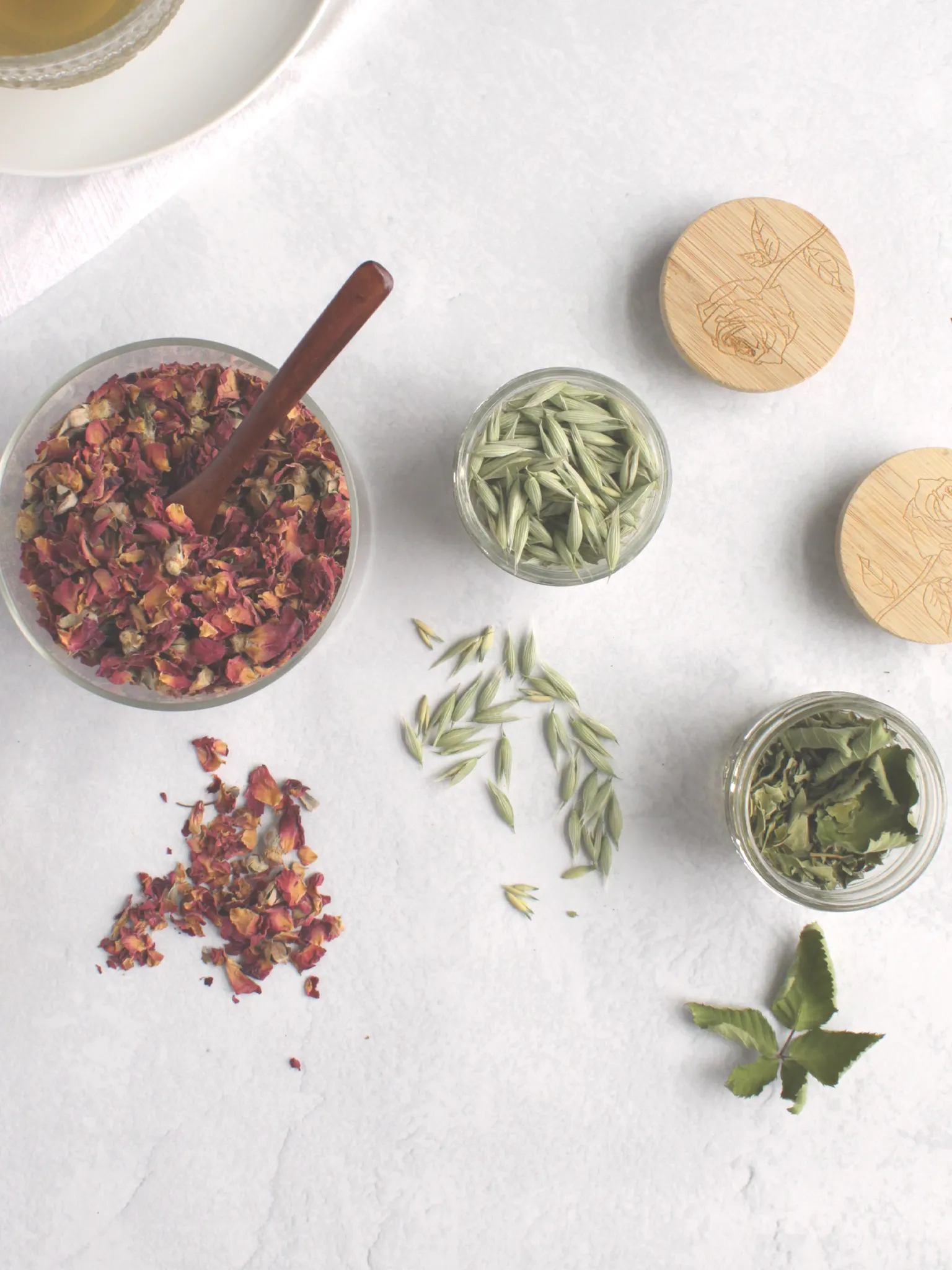 Dried roses, oat tops, and blackberry leaves in glass containers on a counter with a cup of rose tea.