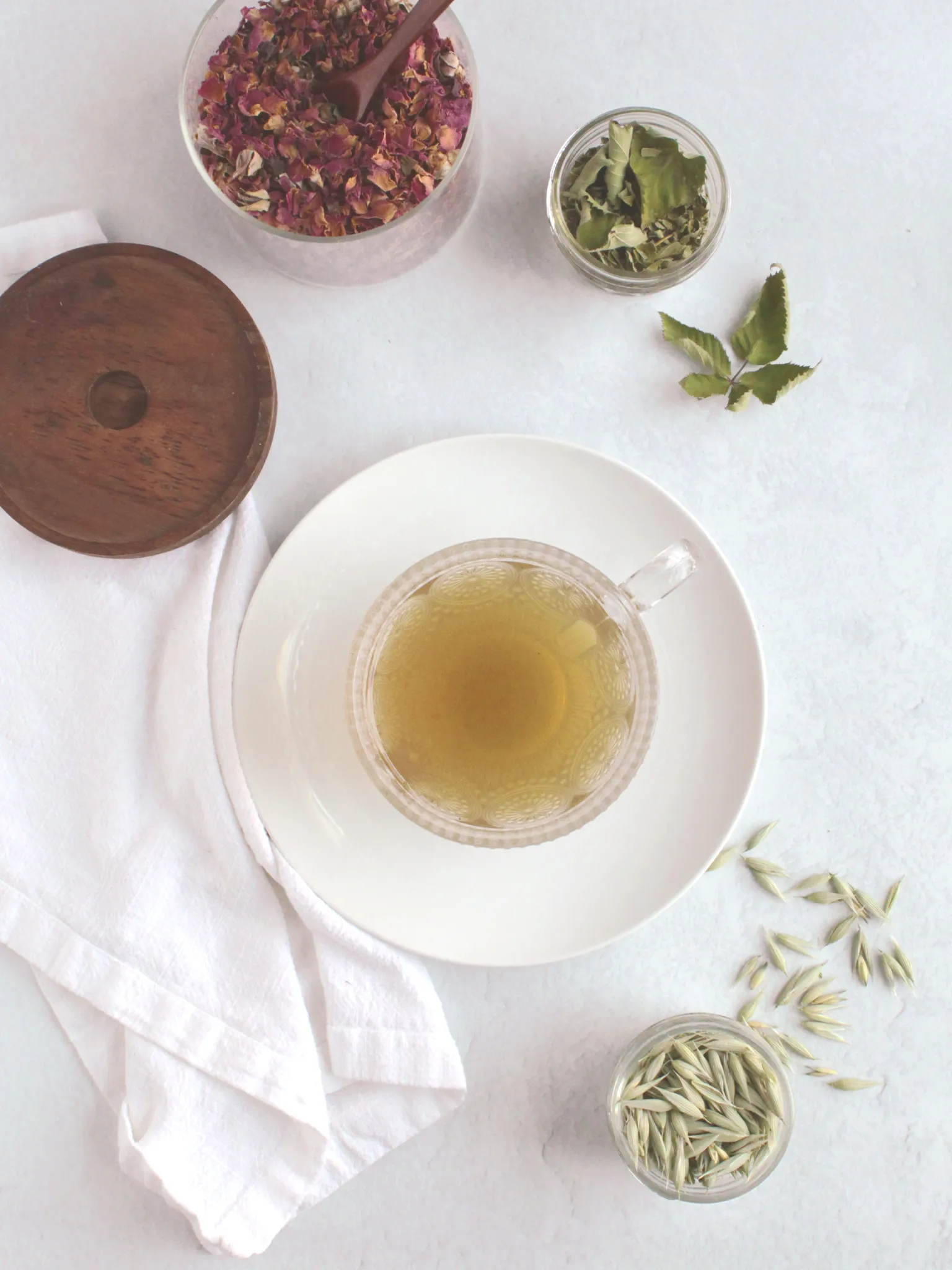 A glass cup of rose blackberry tea on a plate surrounded by containers of dried roses, backberry leaves, and oat tops.