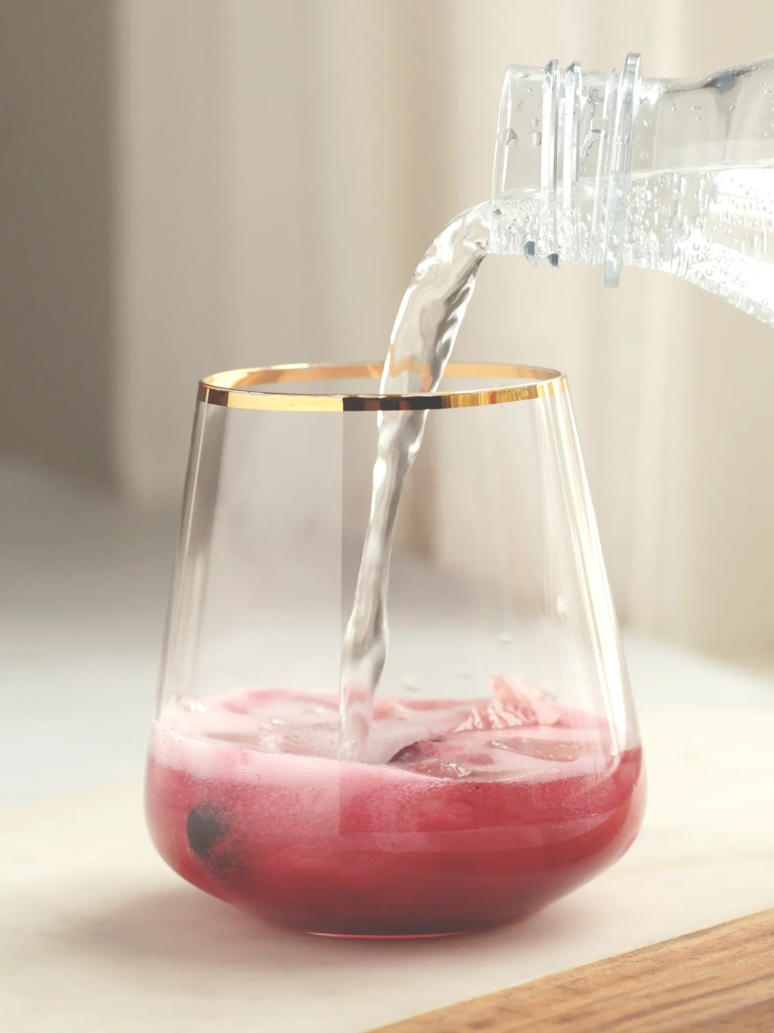 A cocktail glass with gold rim containing frozen blueberries, heart-shaped ice cubes, and blueberry lavender shrub. A hand is pouring club soda into the blueberry lavender shrub.