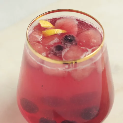 A cocktail glass with gold rim containing blueberry lavender mocktail. There are blueberries and a lemon peel as garnish.