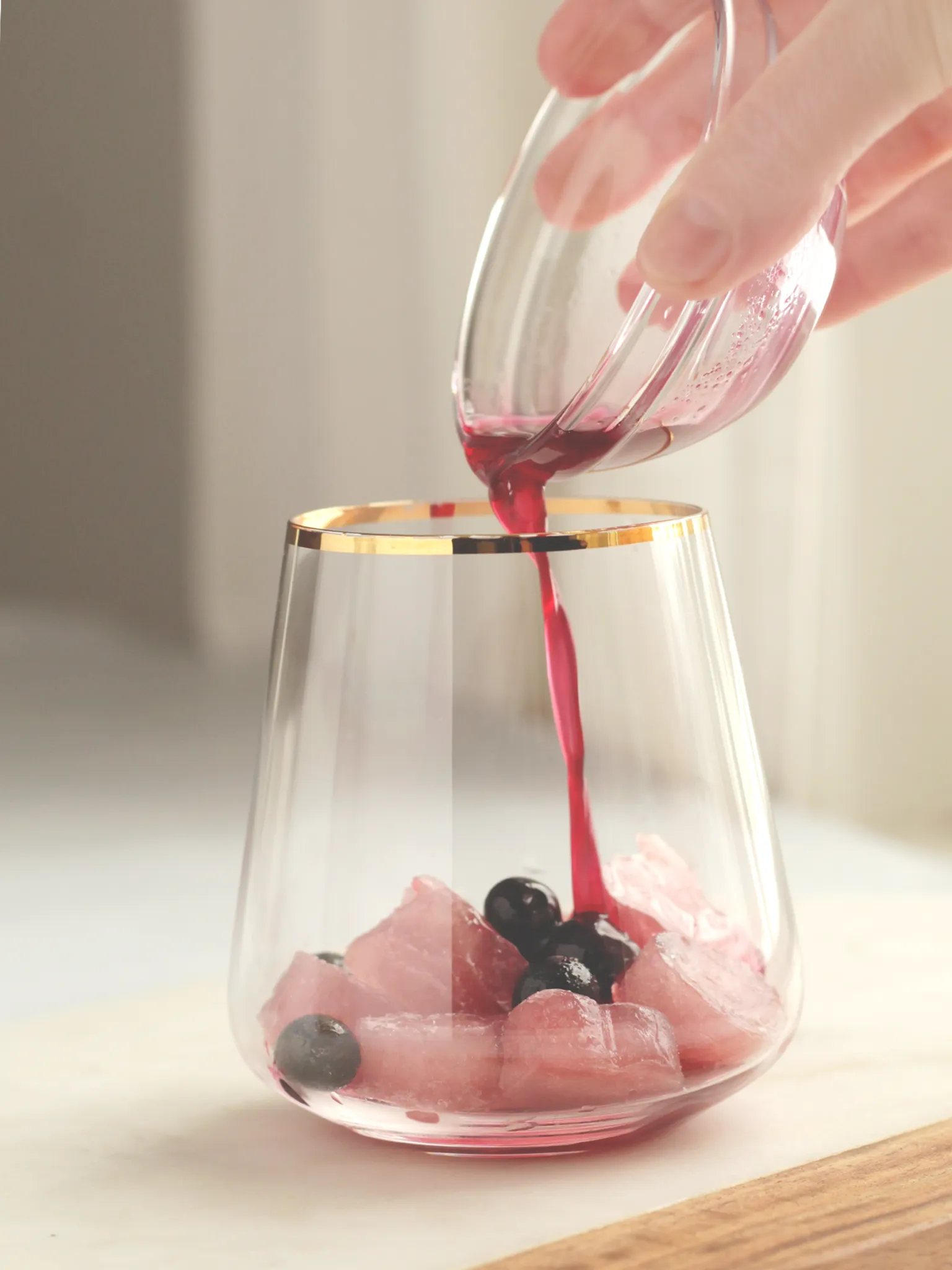 A cocktail glass with gold rim containing frozen blueberries and heart-shaped ice cubes. A hand is pouring blueberry lavender shrub into the glass.