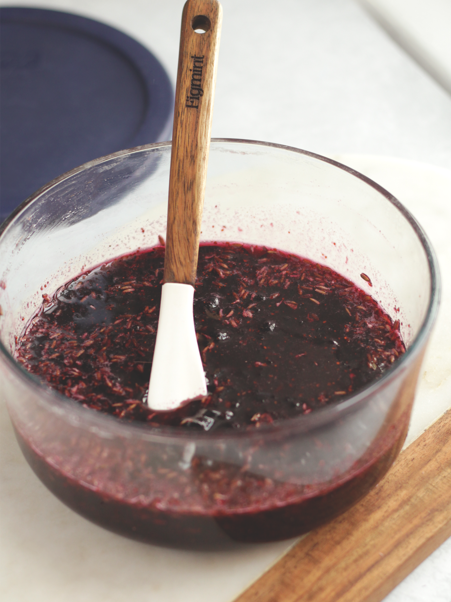 A clear bowl containing mashed blueberries, sugar, and lavender flowers. Everything is mashed together and very liquidy.