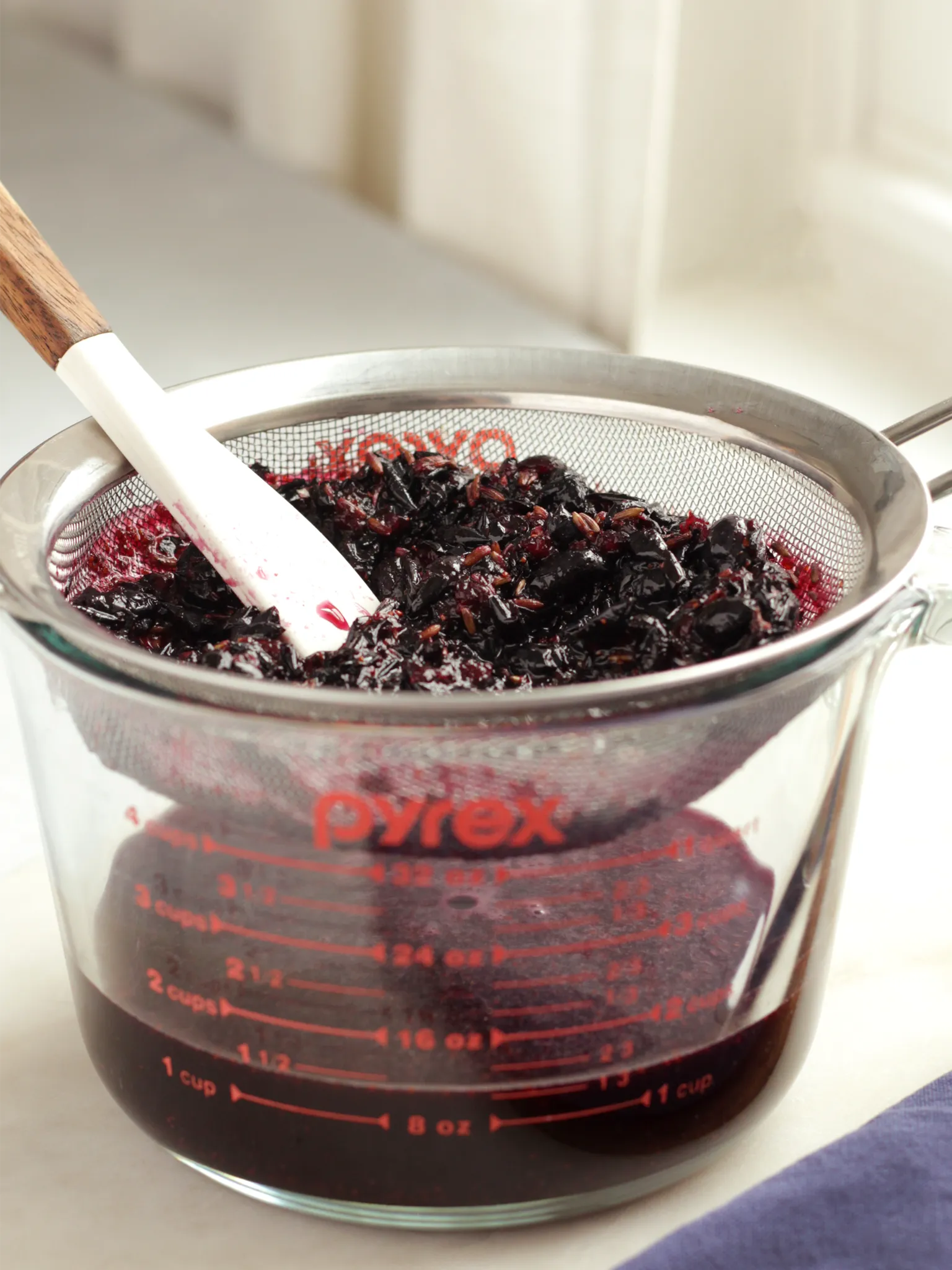 A 4-cup measuring cup with a fine-mesh strainer. The strainer contains the leftover solids from the blueberry, lavender, and sugar mixture. The measuring cup holds the syrup. There is about 1.5 cups of syrup in the measuring cup.