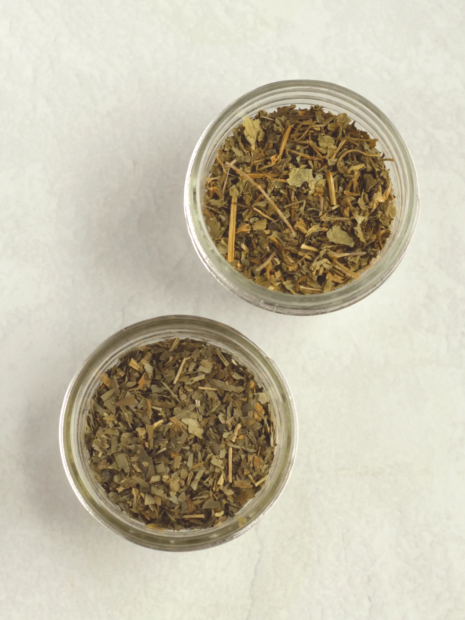 An overhead view of two glass jars filled with dried gotu kola and dried ginkgo leaves.