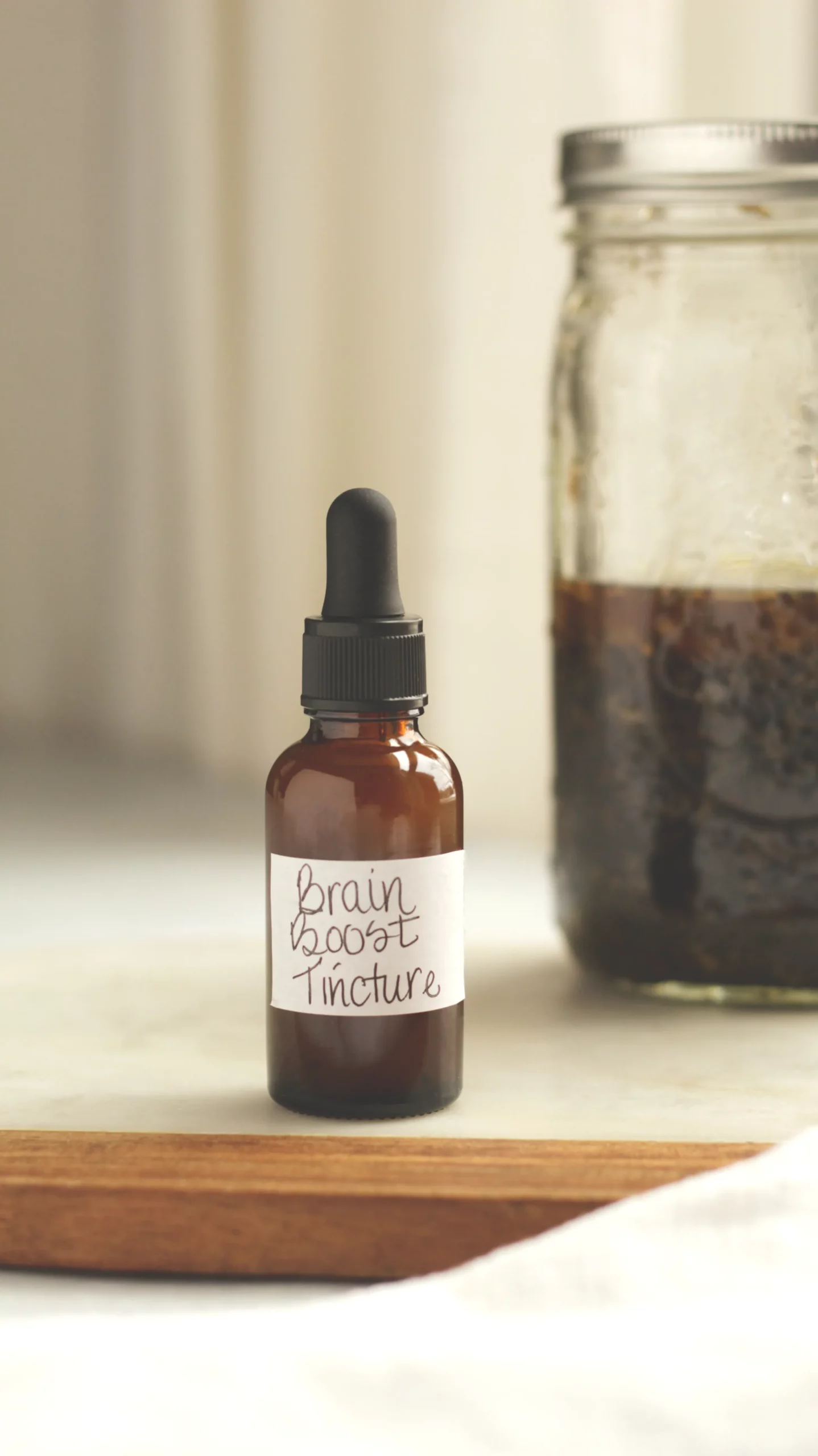 An amber glass dropper bottle labeled 'Brain Booster Tincture' next to a Mason jar filled with dried gotu kola leaves, dried ginkgo leaves and 100 proof vodka. 