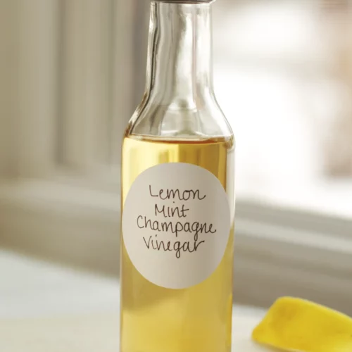 A clear bottle of lemon mint champagne vinegar on a white table next to a lemon peel.