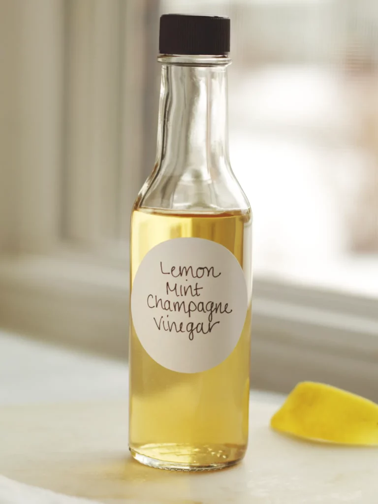 A clear bottle of lemon mint champagne vinegar on a white table next to a lemon peel.