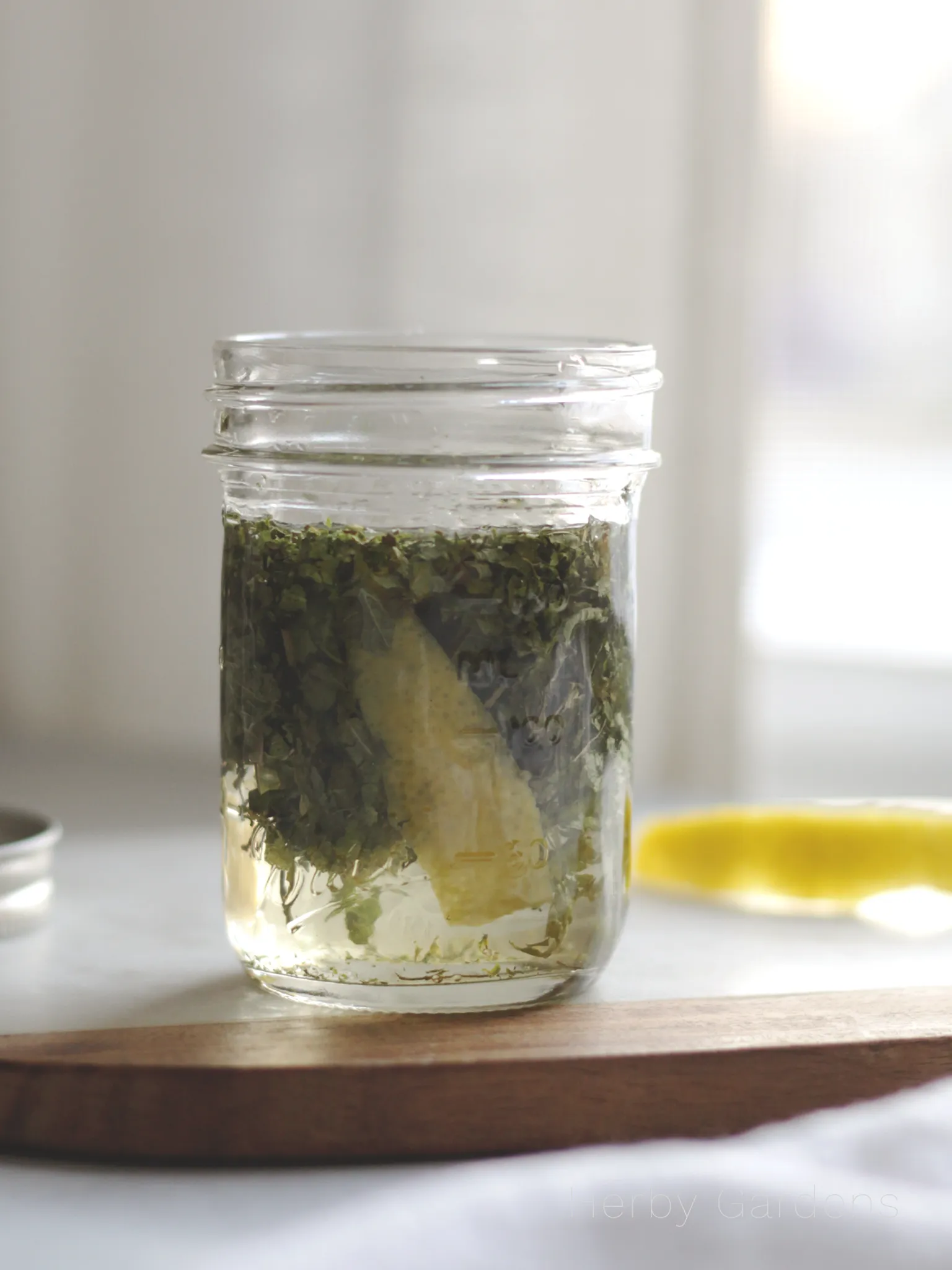 A Mason jar filled with Champagne vinegar, lemon verbena, sweet mint, and a lemon peel. A lemon peel rests on a table in the background.