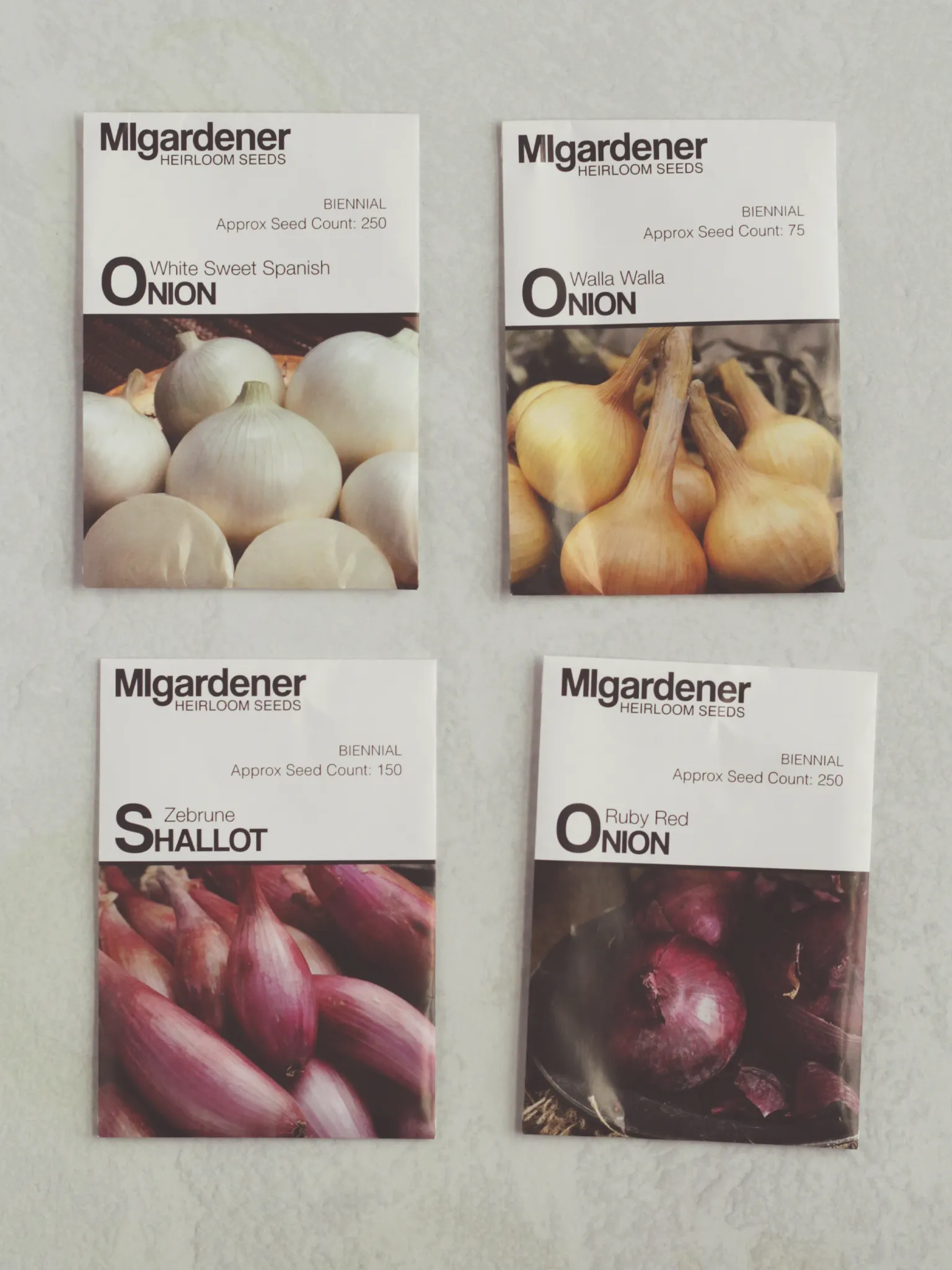 4 seed packets from MIgardener on a table. The seeds are for white sweet Spanish onions, walla walla onions, Zebune shallots, and ruby red onions.