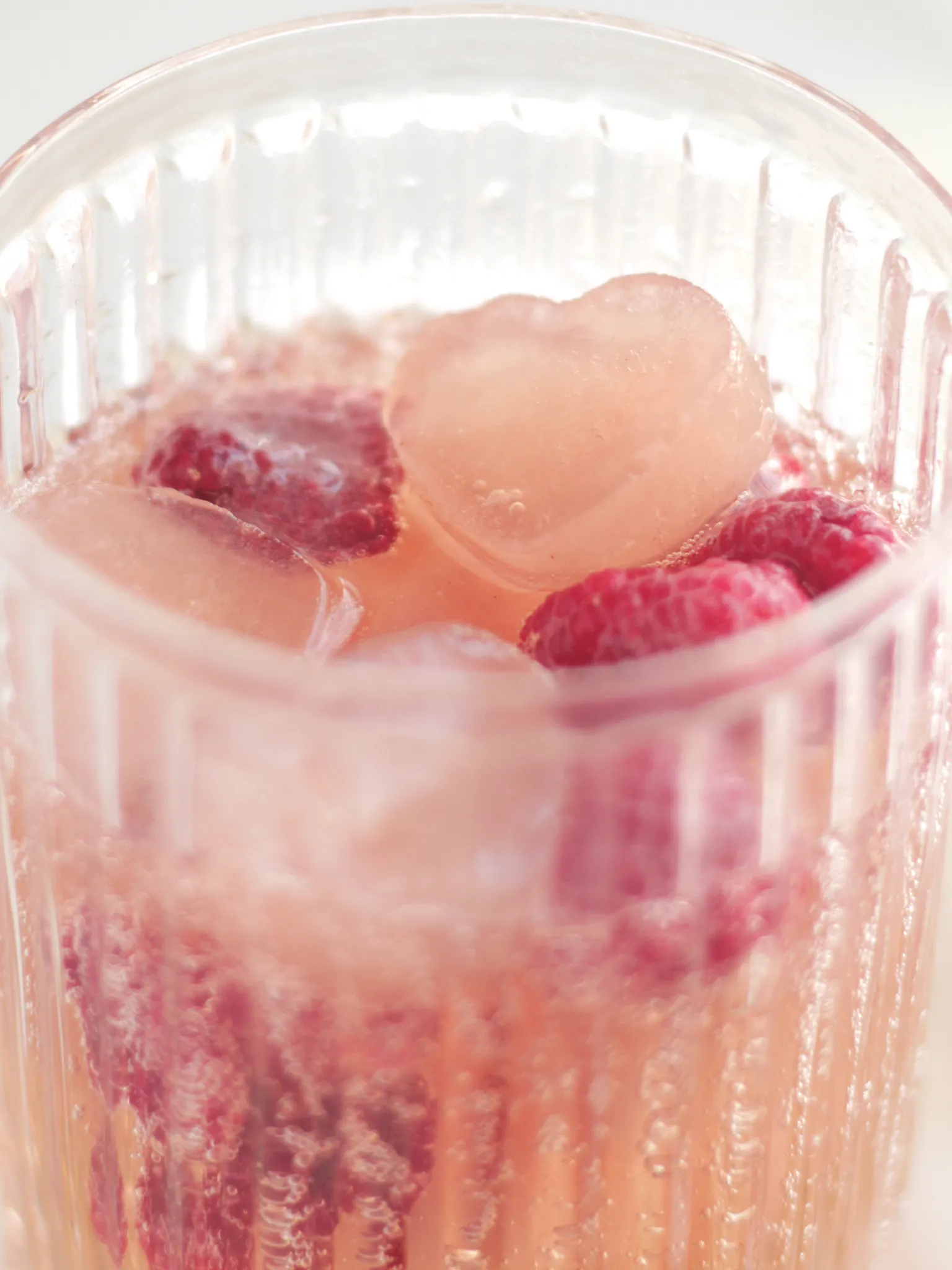 A glass filled with heart shaped ice cubes, frozen raspberries and a fizzy drink.