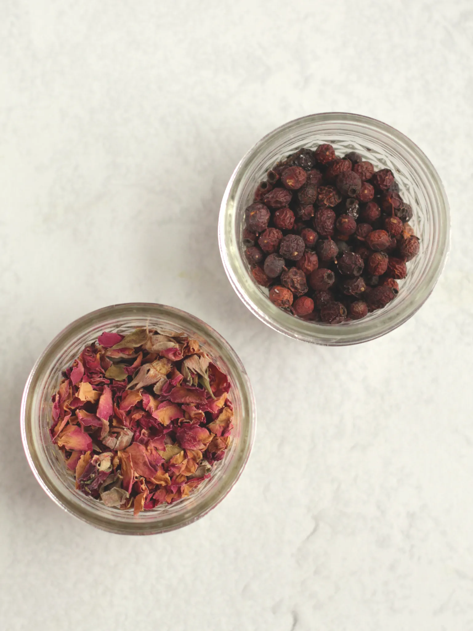 Two small glass jars, filled with dried rose petals, and dried hawthorn berries.