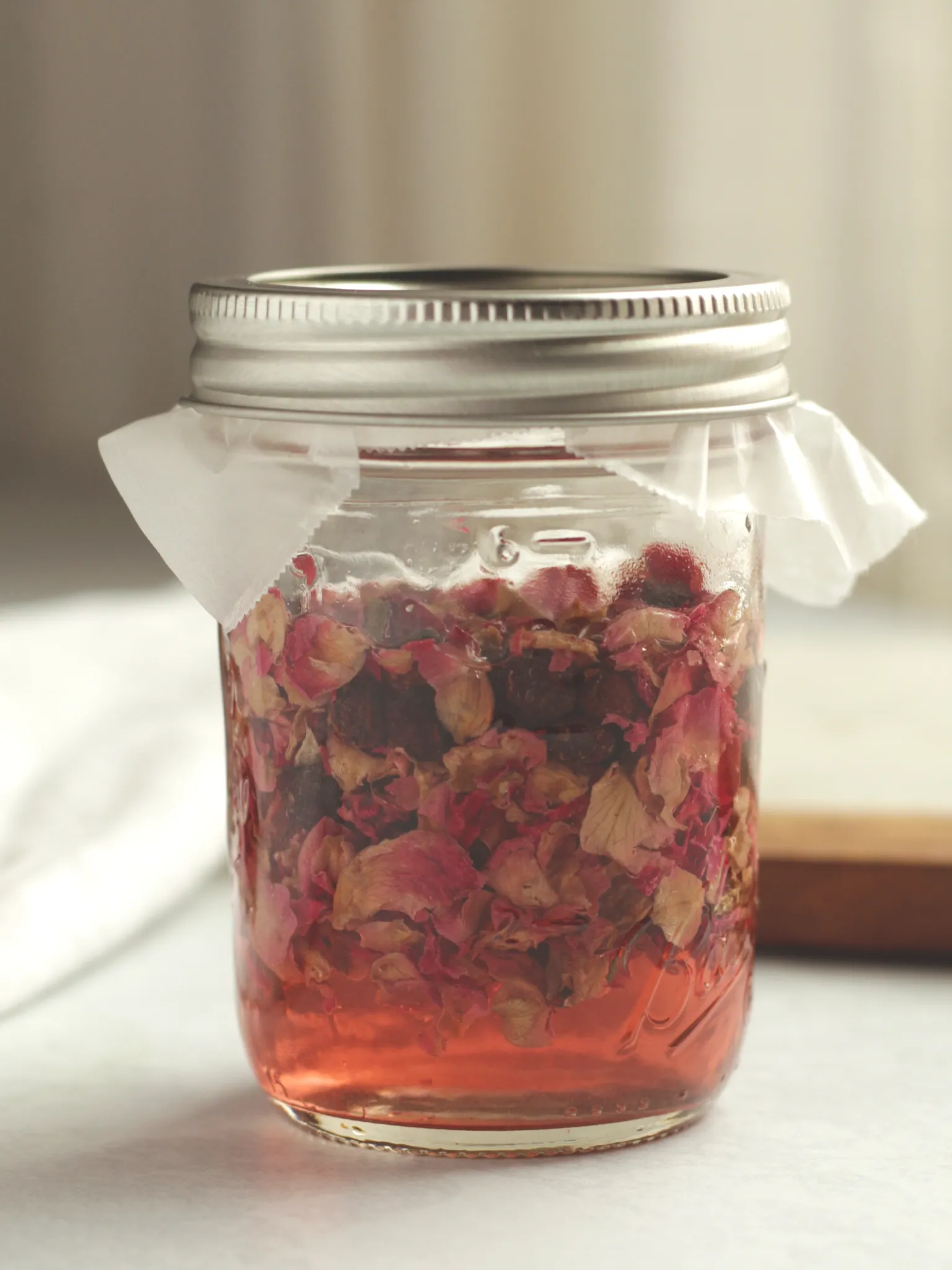 A Mason jar filled with Champagne vinegar, dried roses, and dried hawthorn berries. The top is lined with wax paper.